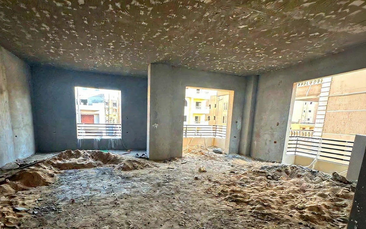 The same room before renovation: bare concrete walls and ceiling, sand on a raw floor, empty window and balcony openings